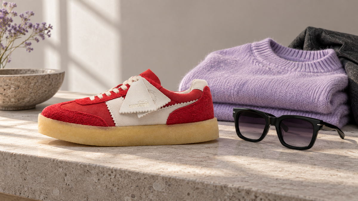 Stilillustration med en röd sneaker, en vikt lavendellila tröja och svarta solglasögon på en ljus stenbänk.